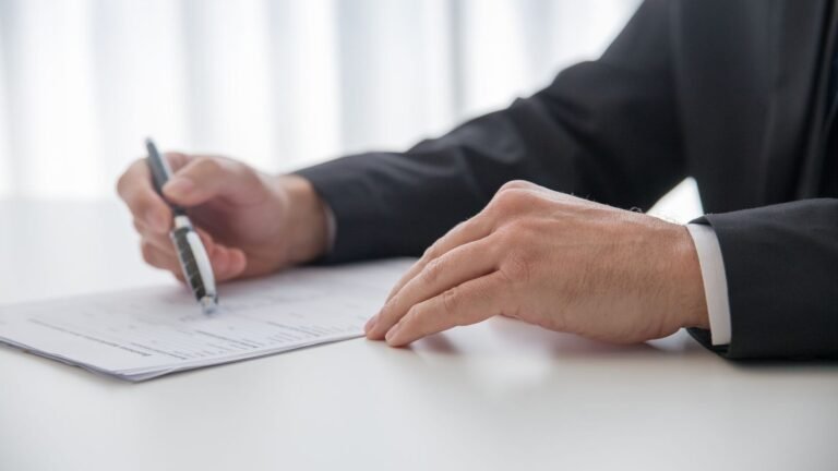 Man in a dark suit signing a document at a white office desk