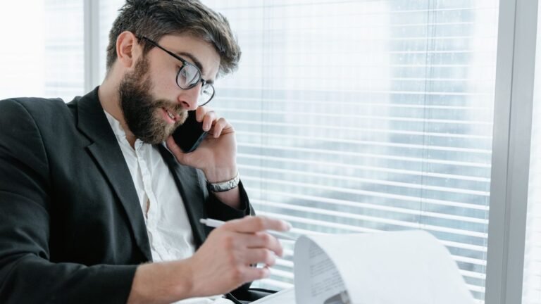 Man on a phone call reviewing papers beside a bright office window