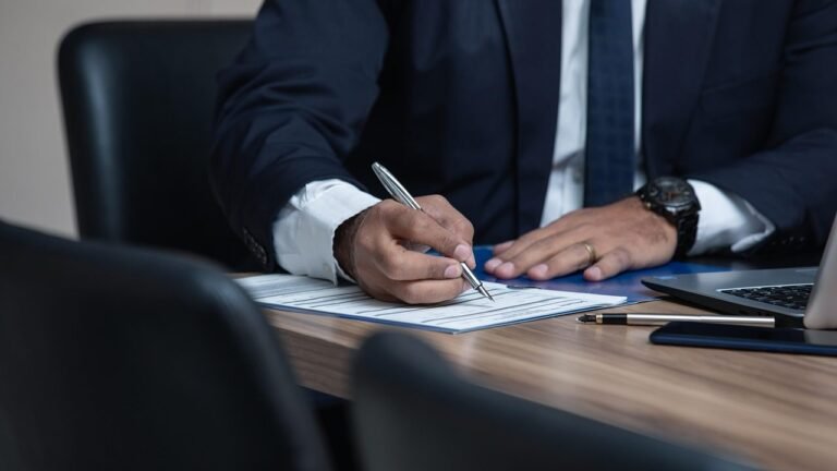 Man in a dark suit signing papers at a desk with a laptop